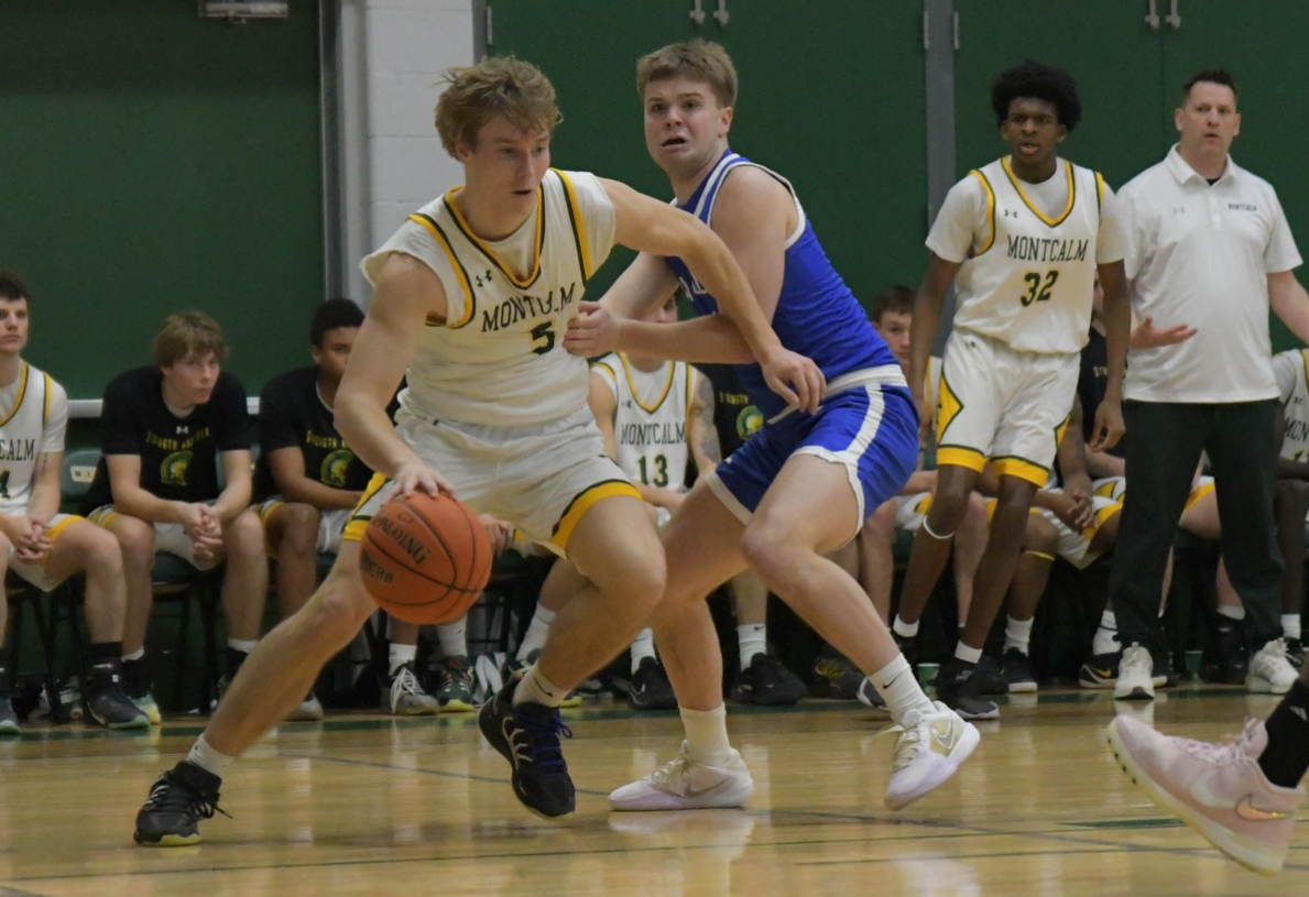 MCC men's basketball player dribbling the ball on the court during a game while oppossing player attempts to block him. He is wearing uniform number 5.