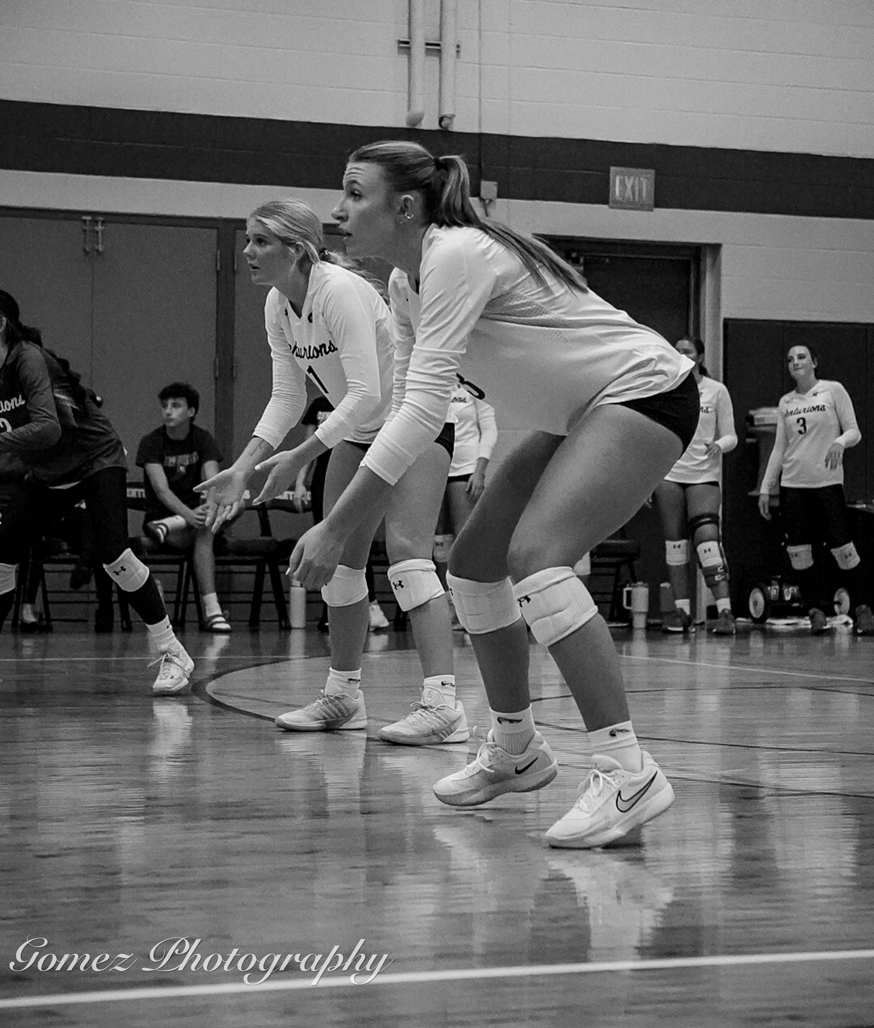 Black and white image of two MCC volleyball players  squatting down and preparing to hit the ball coming over the net. 