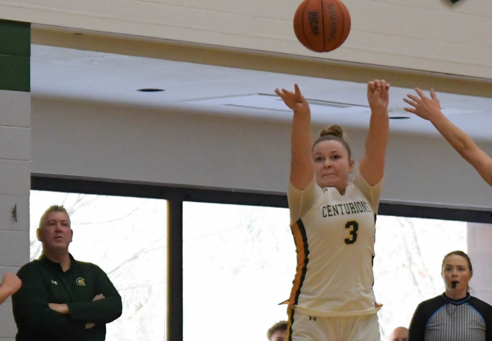 Photo of MCC women's basketball player jumping to shoot the ball while on the court. Coach and players can be seen in the background.
