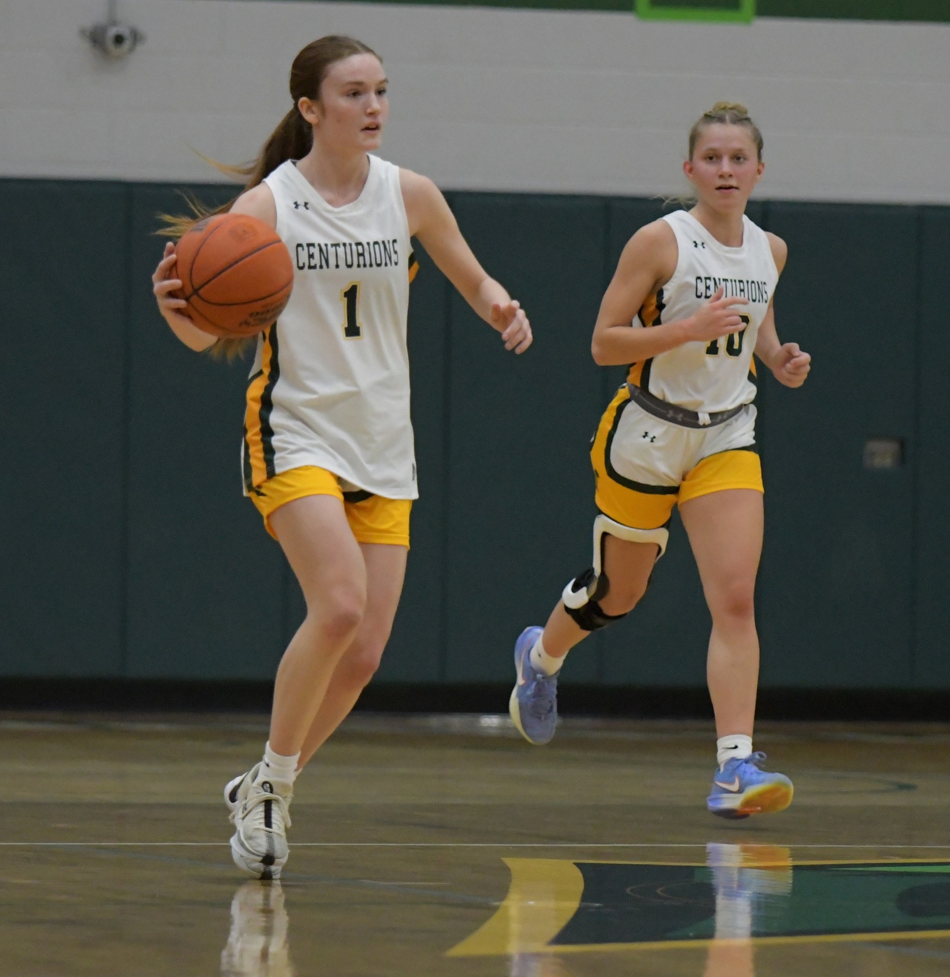 Two MCC women's basketball players on the court during a game. The player wearing number 1 is dribbling the ball while the player wearing the number 10 is running down the court next to her.