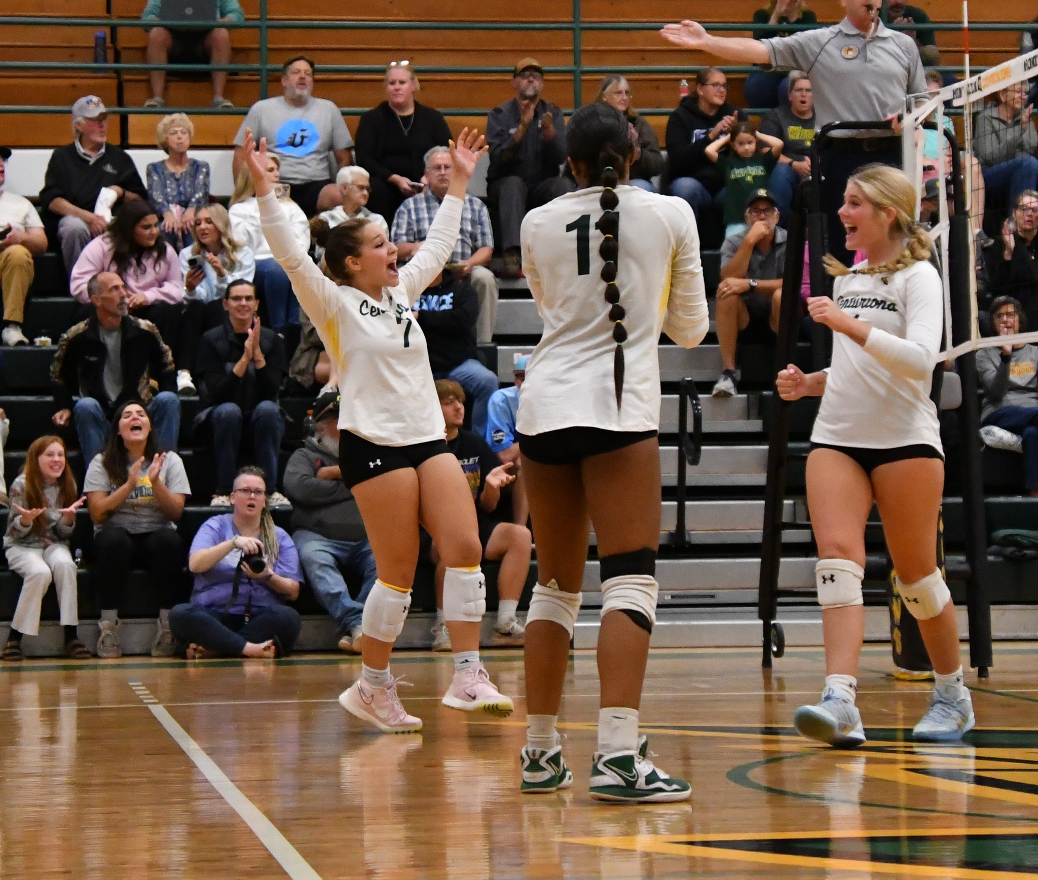 Three Centurion volleyball players celebrate together with arms in the air on the court. 